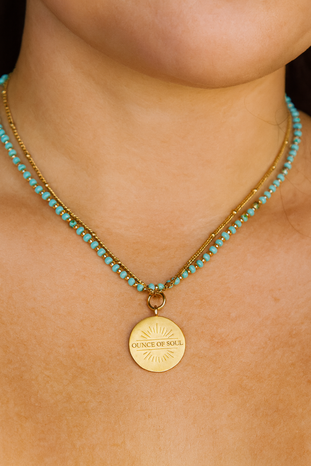 Necklace with turquoise beads and a gold pendant necklace with ounce of soul logo on a person's neck